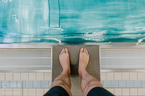 Person steht auf einem Startblock bereit zum Sprung ins Wasser