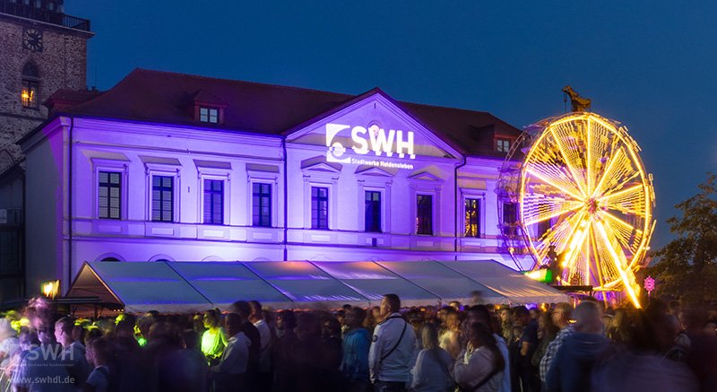 Rathaus Haldensleben zum Altstadtfest im Dunkeln angeleuchtet in violett mit dem SWH-Logo, davor ein Riesenrad