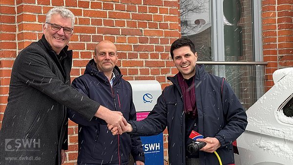 3 Herren geben sich die Hand vor einer Ladesäule