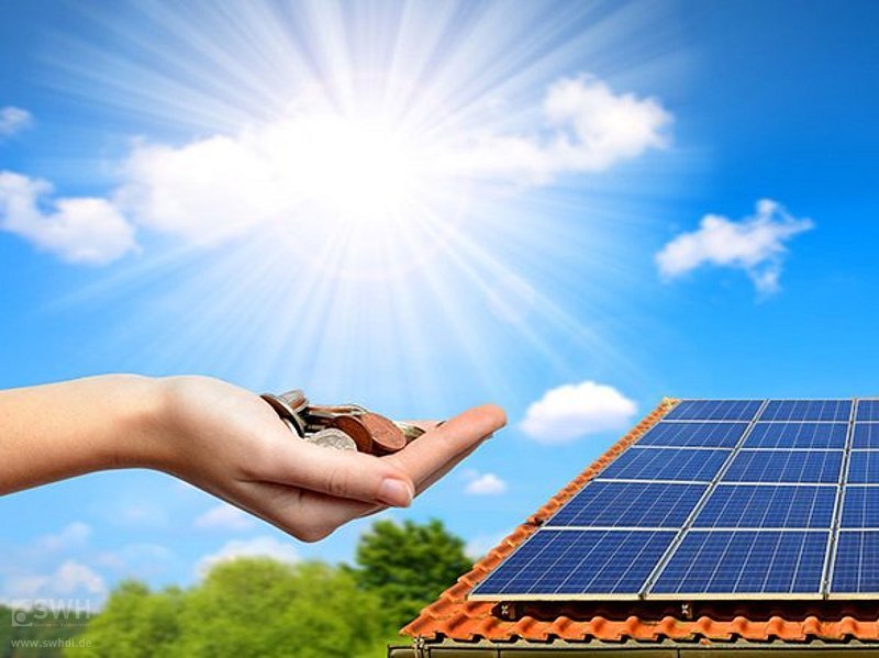 Sonne scheint auf ein Dach mit Photovoltaikanlage, eine Hand daneben mit Münzen